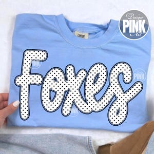 May include: Light blue sweatshirt with the word "Foxes" in a white, polka-dotted font. The word is outlined in black. The sweatshirt has a crew neck and a tag that says "Comfort". A "Design Pink Art" logo is in the upper right corner.