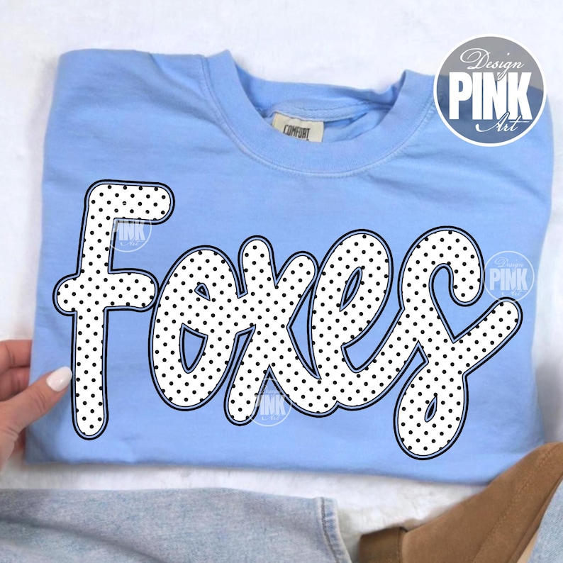 May include: Light blue sweatshirt with the word "Foxes" in a white, polka-dotted font. The word is outlined in black. The sweatshirt has a crew neck and a tag that says "Comfort". A "Design Pink Art" logo is in the upper right corner.