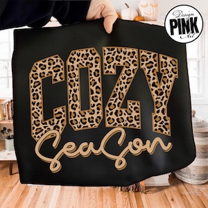 May include: A black blanket featuring the text "COZY Season" in a leopard print pattern. The word "COZY" is in bold, block letters, and "Season" is in a cursive font. The blanket is held up by a person.