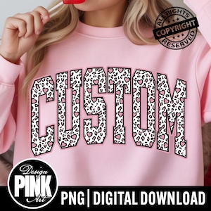 May include: A light pink sweatshirt with the word "CUSTOM" in a leopard print design with pink hearts. The sweatshirt has a crew neck and long sleeves. The image also includes the text "PNG | DIGITAL DOWNLOAD".