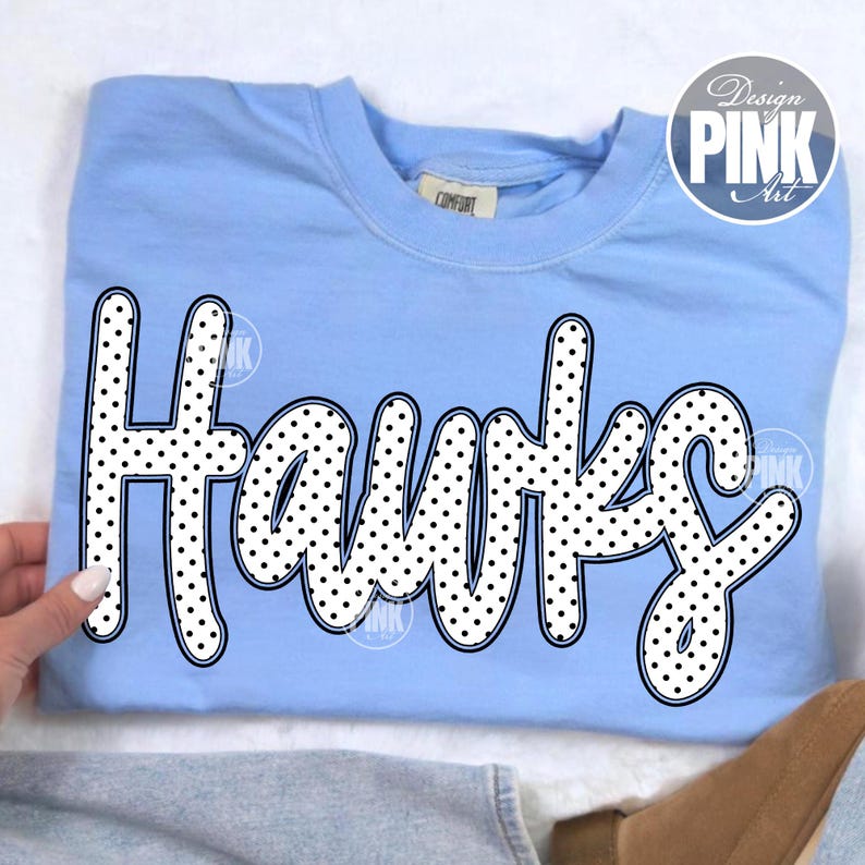 May include: A light blue sweatshirt with the word "Hawks" in large, white, polka-dotted letters. The word is outlined in black. The sweatshirt has a crew neck and a tag that says "Comfort". The design is by Design Pink Art.