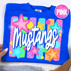 May include: A royal blue sweatshirt with the word "Mustangs" in white script. The design includes colorful stars, circles, and vertical bars in pink, yellow, green, and orange. The design is labeled "Design PINK Art".