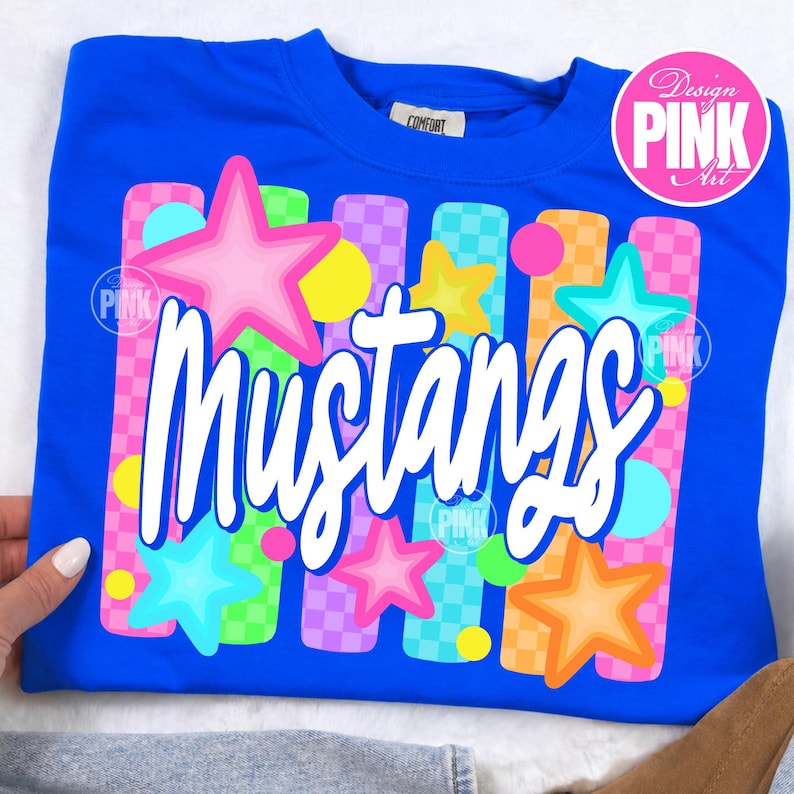 May include: A royal blue sweatshirt with the word "Mustangs" in white script. The design includes colorful stars, circles, and vertical bars in pink, yellow, green, and orange. The design is labeled "Design PINK Art".