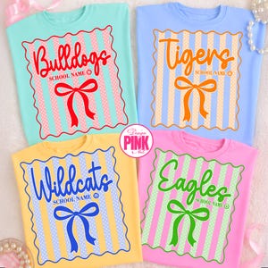 May include: Four colorful t-shirts with school mascot designs. Each shirt features a team name in a script font, a bow, and vertical stripes. The shirts are light blue, yellow, pink, and blue.
