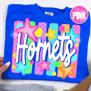May include: A royal blue sweatshirt with the word "Hornets" in white script. The design includes colorful stars, circles, and vertical stripes in pink, yellow, green, and orange. The shirt has a "Comfort" tag.