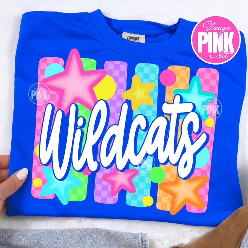May include: A royal blue sweatshirt featuring the word "Wildcats" in white script. The design includes colorful stars, circles, and vertical stripes in pink, yellow, orange, and blue. The sweatshirt has a crew neck and a small tag that says "Comfort".