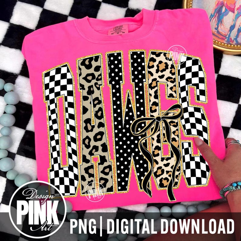 May include: A bright pink sweatshirt with the word "DAWGS" in a bold, decorative font. The letters feature a mix of patterns, including checkered, leopard print, and polka dots, with gold trim. A black bow is centered below the letters.