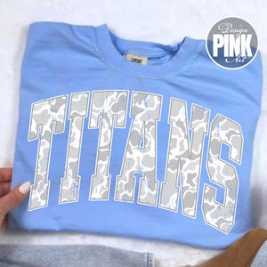 May include: A light blue crew neck t-shirt featuring the word TITANS in large, arched block letters. The lettering has a grey and white camouflage pattern with a distressed, weathered texture. A small tag is visible at the inner neckline.