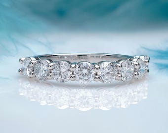 May include: A silver ring with seven round, clear gemstones set in a row. The ring is displayed on a reflective white surface with a blurred blue background, creating a soft, elegant aesthetic. The ring's design is simple and classic.