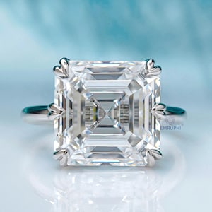 May include: A square-cut diamond ring with a silver band. The large, clear diamond is the focal point, set with four decorative prongs. The ring is displayed against a light blue and white background.