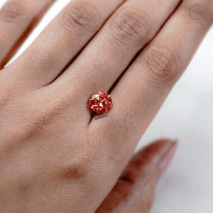 Puede incluir: Una gema redonda, facetada, de color naranja rojizo. La gema se sostiene en una mano, con el texto "EMRUPHI" visible en la gema. La gema tiene un corte brillante, que refleja la luz.
