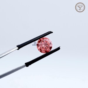 Puede incluir: Un diamante rosa redondo y facetado es sostenido por unas pinzas. El diamante es el punto focal, mostrando su talla y color. El fondo es blanco y neutro. El texto "EMRUP" es visible.