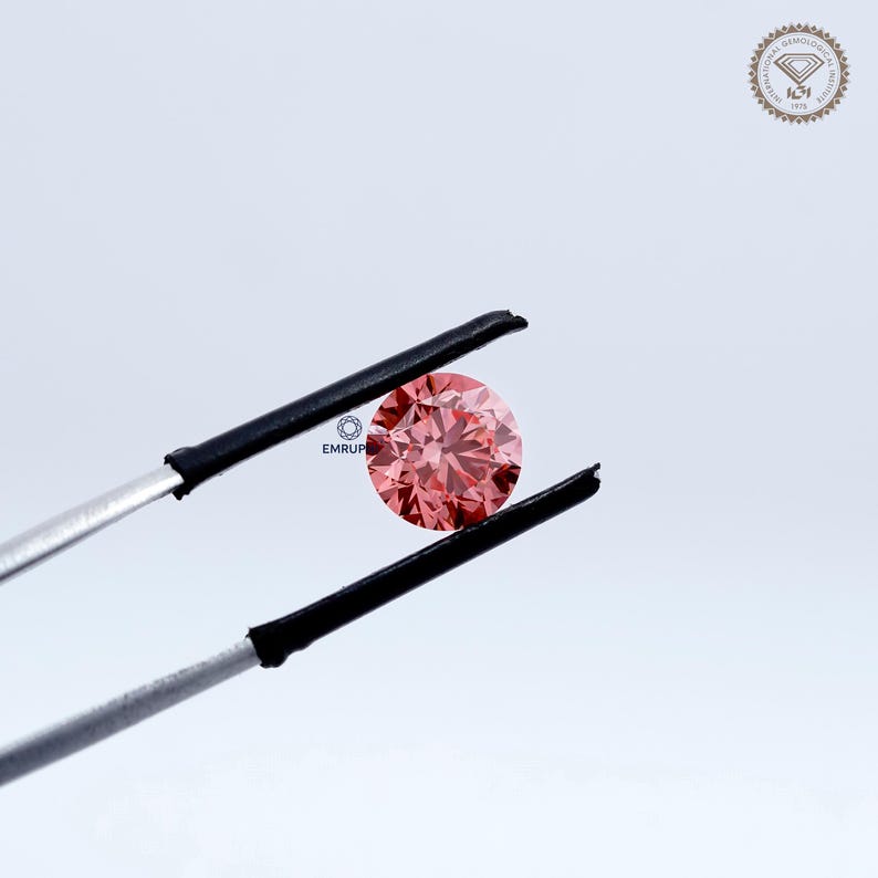 Puede incluir: Un diamante rosa redondo y facetado es sostenido por unas pinzas. El diamante es el punto focal, mostrando su talla y color. El fondo es blanco y neutro. El texto "EMRUP" es visible.