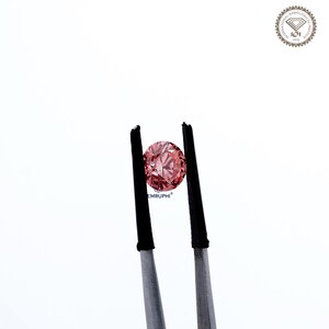 Puede incluir: Un diamante rosa redondo es sostenido por unas pinzas con puntas negras sobre un fondo blanco. El diamante tiene m&uacute;ltiples facetas que reflejan la luz. La imagen incluye el texto "EMRUPHI" y un logotipo circular.