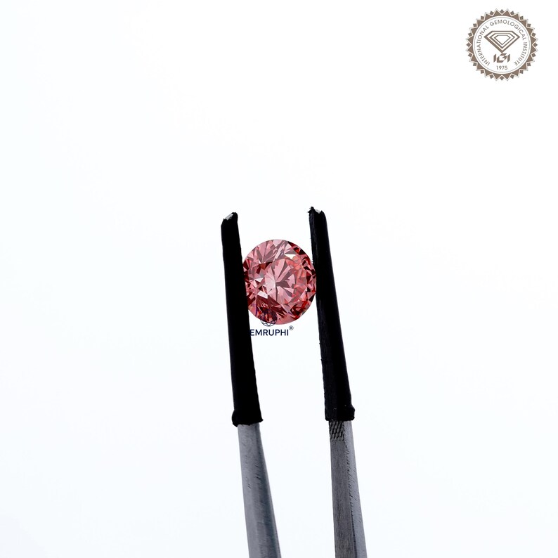 Puede incluir: Un diamante rosa redondo es sostenido por unas pinzas con puntas negras sobre un fondo blanco. El diamante tiene m&uacute;ltiples facetas que reflejan la luz. La imagen incluye el texto "EMRUPHI" y un logotipo circular.
