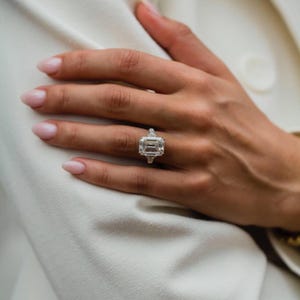 May include: An emerald-cut diamond ring with two side stones. The ring is set on a hand with manicured nails, against a white background. The ring's large center stone is rectangular, flanked by smaller, triangular stones.