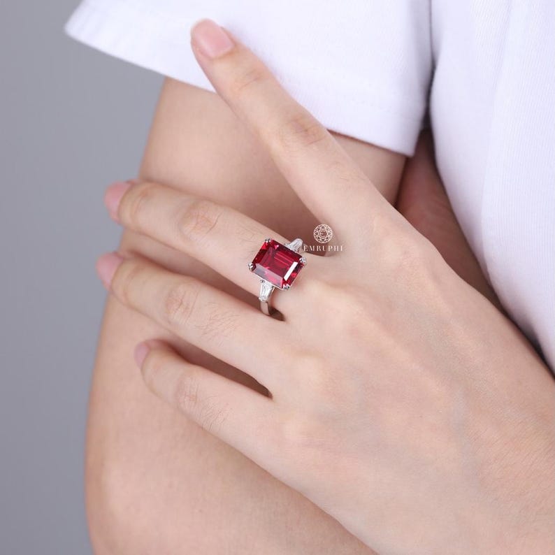 Pode incluir: Um anel de prata com uma grande pedra retangular vermelha escura. Duas pedras claras triangulares menores ladeiam a pedra principal. O anel est&aacute; em um dedo, mostrando seu design e cor.