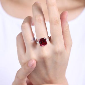 Pode incluir: Um anel de prata com uma grande pedra retangular vermelha escura como pe&ccedil;a central. Duas pedras menores e claras ladeiam a pedra principal. O anel &eacute; exibido em uma m&atilde;o, mostrando seu design e brilho.