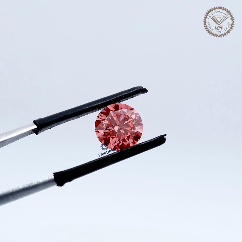 Puede incluir: Un diamante rosa redondo y facetado, sostenido por unas pinzas con puntas negras. La gema tiene facetas visibles que reflejan la luz. El fondo es blanco, con un logotipo en la esquina superior derecha.