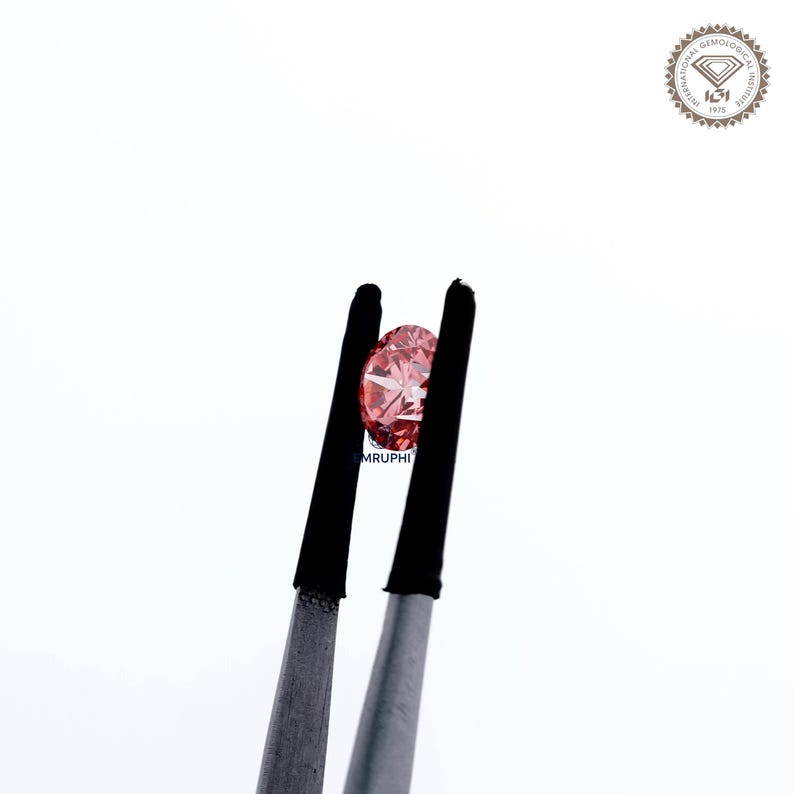 Puede incluir: Un diamante rosa claro tallado en forma ovalada es sujetado por unas pinzas. Las facetas del diamante reflejan la luz. Las pinzas tienen puntas de goma negra. El fondo es blanco. El texto "MRUPHI" es visible.