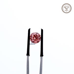 Puede incluir: Un diamante rosa redondo y facetado sostenido por unas pinzas con puntas negras. El diamante es el punto focal sobre un fondo blanco. El logotipo del Instituto Gemol&oacute;gico Internacional est&aacute; en la esquina superior derecha.
