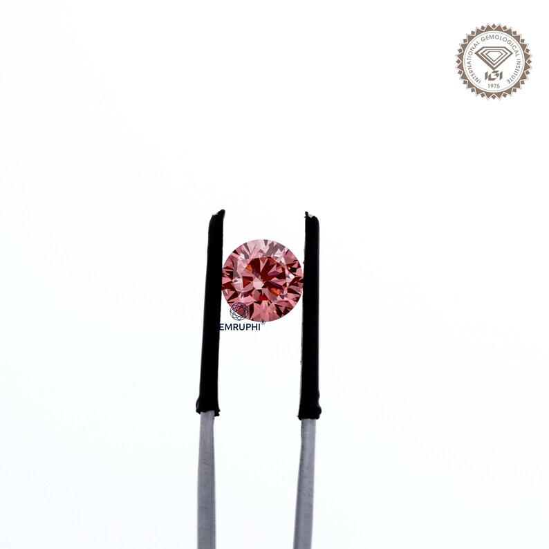 Puede incluir: Un diamante rosa redondo y facetado sostenido por unas pinzas con puntas negras. El diamante es el punto focal sobre un fondo blanco. El logotipo del Instituto Gemol&oacute;gico Internacional est&aacute; en la esquina superior derecha.