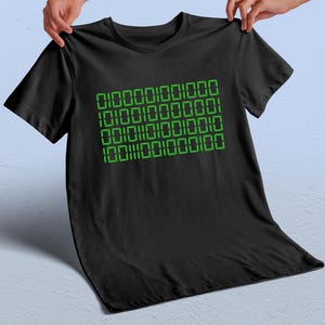 May include: Black t-shirt with a green binary code design. The binary code is arranged in four rows of numbers. The t-shirt is made of a soft material.