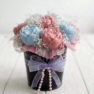 May include: A bouquet of pink and blue peony-shaped soaps in a black bucket. The bucket is decorated with a lavender ribbon and a string of pearls. The arrangement includes baby's breath and is set on a white wooden surface.