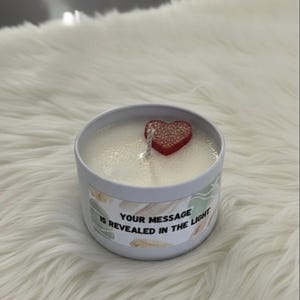 May include: A white candle in a silver tin with a red heart-shaped decoration. The candle has a wick and the text "YOUR MESSAGE IS REVEALED IN THE LIGHT" is printed on the side. The candle sits on a white, fluffy surface.