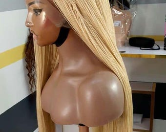 May include: A blonde braided wig on a mannequin head. The wig is long and straight, with a natural-looking hairline. The mannequin is on a stand, and other wigs are visible in the background.