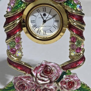 May include: A decorative gold-toned clock with a white face and Roman numerals. The clock is adorned with pink roses, red ribbon accents, and colorful gemstones. The clock face reads "JANESEYMOUR" and "St.Catherine's Court".
