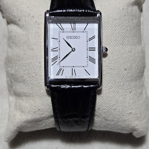 May include: A rectangular Seiko wristwatch with a silver-tone case, a white dial featuring black Roman numeral hour markers, and black hands. The watch is fitted with a black textured leather strap and includes a small crown on the right side.
