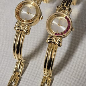 May include: Two gold-toned wristwatches with matching bands. The watch faces are silver with gold hands and Roman numerals. One watch has a bezel of orange-colored stones, the other has a bezel of red stones. The bands have a bow design.