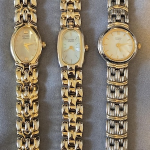 May include: Three gold-toned and silver-toned Citizen watches with different band designs. The watch faces are light-colored with gold accents. The watch bands have a variety of link styles. The watches are displayed on a gray surface.