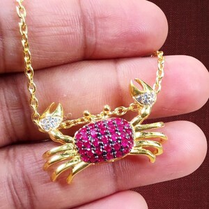 Può includere: Una collana con pendente a forma di granchio dorato. Il corpo del granchio è incastonato con piccole gemme rosse rotonde. Gli artigli hanno accenti chiari e scintillanti. Anche la catena della collana è dorata.