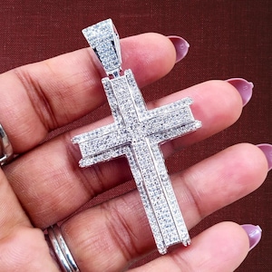 May include: A silver-colored cross pendant encrusted with numerous small, clear gemstones. The cross has a rectangular bale for a chain and is held against a brown background.