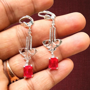 Puede incluir: Un par de pendientes colgantes plateados con una gema roja rectangular en la parte inferior. Los pendientes tienen un diseño Art Déco con una parte superior decorativa y un cierre de palanca. Los pendientes se sostienen en una mano.