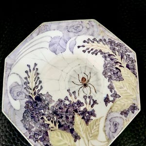 Op de afbeelding: Achthoekige decoratieve plaat met een spinnenwebontwerp. De plaat heeft een spin in het midden, omringd door paarse bloemen en groene bladeren. De randen van de plaat zijn lichtpaars.