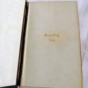 May include: An open antique book with a cream-colored page featuring the gold text "Anna Kot." and "1888." The book's spine is black, and the left page has a white, patterned design.