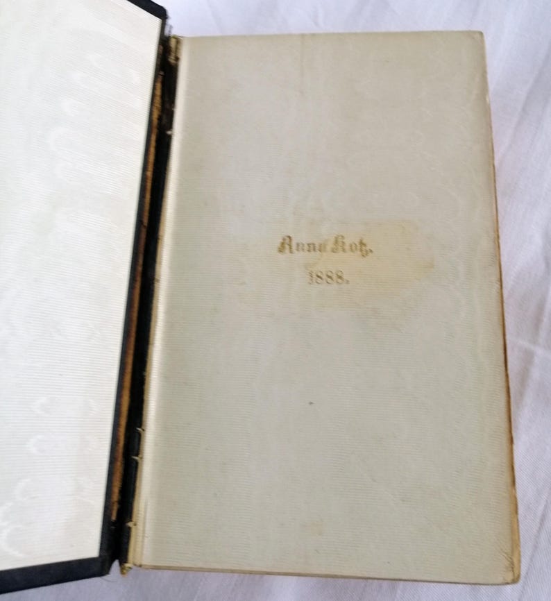 May include: An open antique book with a cream-colored page featuring the gold text "Anna Kot." and "1888." The book's spine is black, and the left page has a white, patterned design.