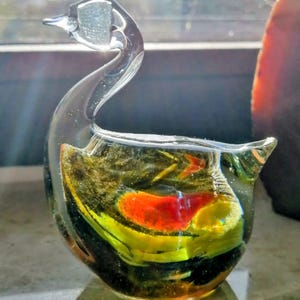 Könnte beinhalten: Eine durchsichtige Glas-Schwan-Figur mit gebogenem Hals und detailliertem Schnabel. Das Innere des Schwans ist mit bunten abstrakten Formen in Rot, Gelb und Grün gefüllt. Die Figur wird auf einer reflektierenden Oberfläche präsentiert.