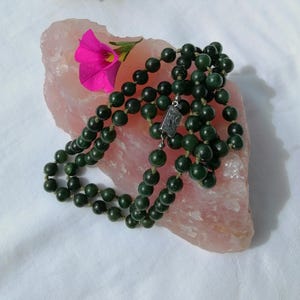 Può includere: Collana di perline verde scuro con chiusura argentata, appoggiata su una pietra rosa chiaro. Un fiore rosa brillante con centro giallo è posto sulla pietra. La collana è fatta di perline rotonde.