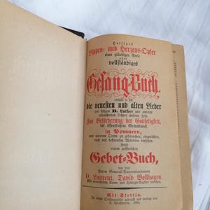 May include: An open antique book with a tan cover and pages. The title page features ornate red and black text in German, including "Gelang-Buch" and "Gebet-Buch." The book appears to be from 1880.