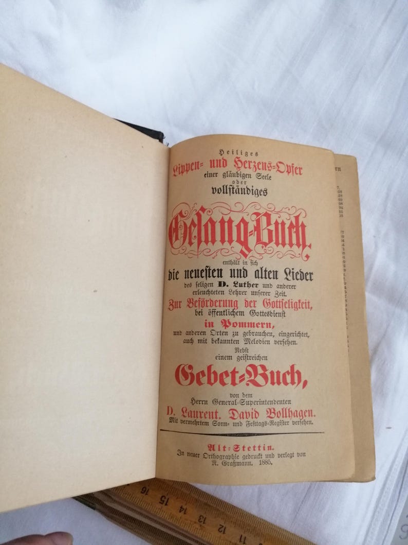 May include: An open antique book with a tan cover and pages. The title page features ornate red and black text in German, including "Gelang-Buch" and "Gebet-Buch." The book appears to be from 1880.