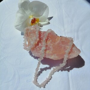 May include: A light pink rose quartz necklace draped over a large, rough-hewn rose quartz crystal. A white orchid flower with a yellow and red center rests nearby. The items are arranged on a white surface.