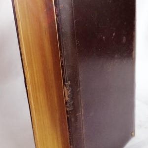 May include: A vintage book with a dark brown leather cover and gold-colored page edges. The book is secured with brass-colored rivets. The pages are aged, and the spine shows signs of wear, suggesting it is an antique.