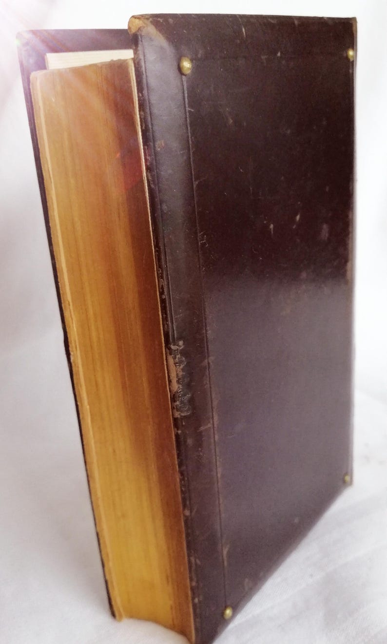 May include: A vintage book with a dark brown leather cover and gold-colored page edges. The book is secured with brass-colored rivets. The pages are aged, and the spine shows signs of wear, suggesting it is an antique.