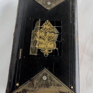 May include: An antique black book with gold-colored metal accents. The book has a decorative gold emblem with text in the center and gold corner pieces. The book is rectangular and has a worn appearance.