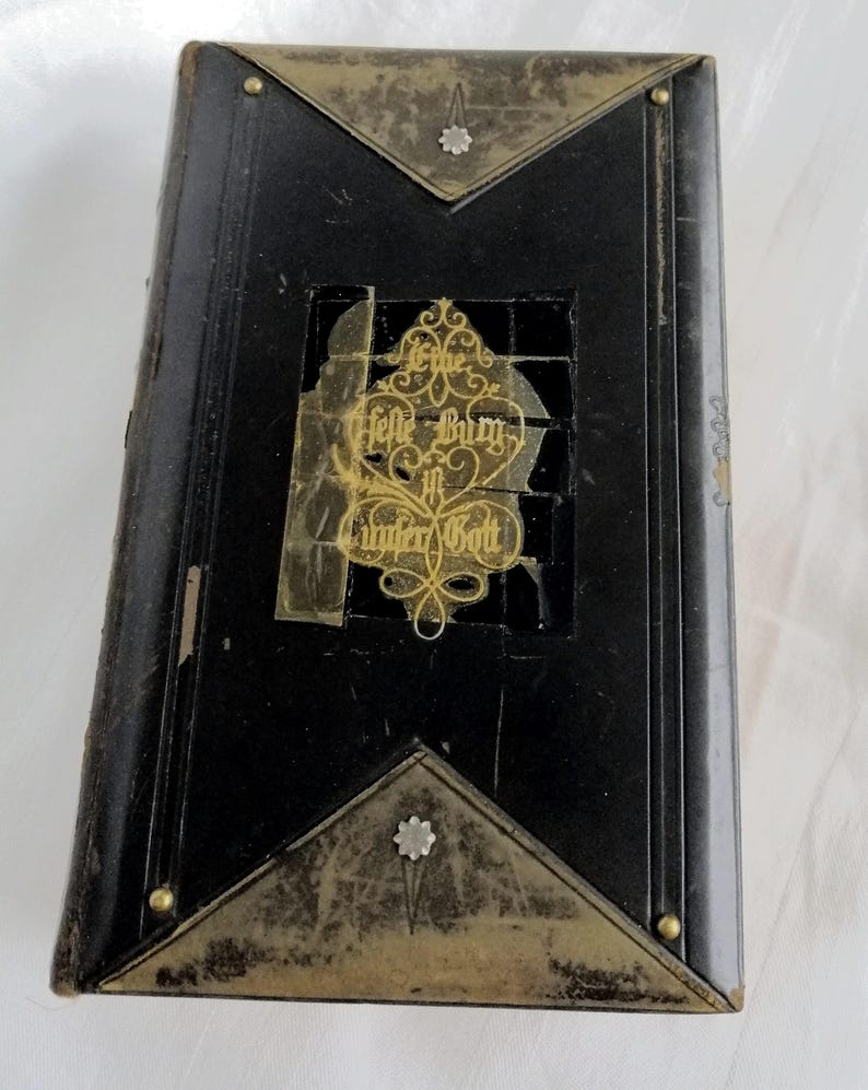 May include: An antique black book with gold-colored metal accents. The book has a decorative gold emblem with text in the center and gold corner pieces. The book is rectangular and has a worn appearance.