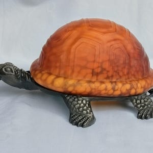 Könnte beinhalten: Eine dekorative Schildkrötenlampe mit einer orange-braunen Glasschale. Die Lampe hat einen dunklen Metallsockel und Kopf, und die Schale ist beleuchtet. Die Lampe ist als Schildkröte gestaltet.